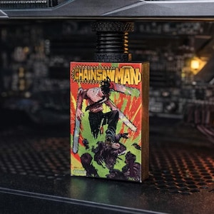 May include: Inside a computer case, a GeForce RTX graphics card is visible alongside a small "Chainsaw Man" comic book. The graphics card is dark grey with green detailing, and the comic book has a colourful, dynamic cover.