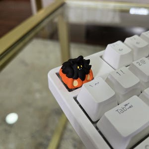 May include: A custom keycap featuring a black creature with yellow eyes and a fluffy appearance, set on a white keyboard. The keycap is orange and black, adding a unique touch to the keyboard.