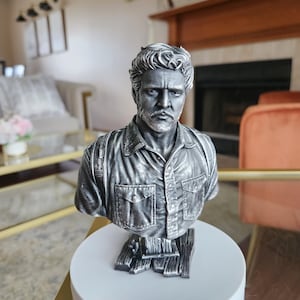 Peut inclure: Buste argenté d'un homme au regard sérieux, vêtu d'une chemise à poches. La sculpture est montée sur une base circulaire blanche, avec des détails de la chevelure, de la barbe et des vêtements. Objet décoratif.