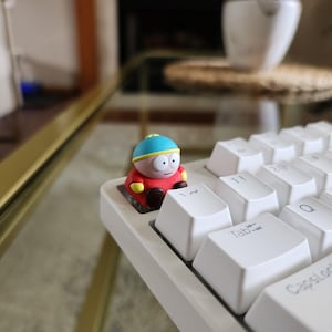 May include: A white mechanical keyboard with a cartoon character figurine perched on a key. The figurine is red, yellow, and blue. The keyboard keys are white with black lettering.