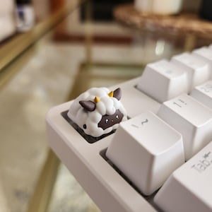 Puede incluir: Una tecla de teclado personalizada con forma de oveja. La tecla tiene un cuerpo blanco y esponjoso, una cara marrón y pequeños cuernos amarillos. La tecla está instalada en un teclado blanco.