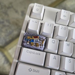 May include: A custom keycap featuring a miniature, detailed weapon design in blue, gold, and red, set against a textured gray background. The keycap is installed on a white mechanical keyboard, with the CapsLock key visible.