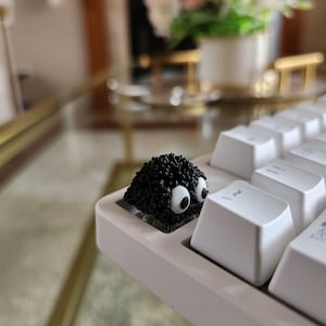 Op de afbeelding: Een wit mechanisch toetsenbord met een aangepaste zwarte keycap in de vorm van een fuzzy wezen met grote witte ogen. Het toetsenbord staat op een glazen tafel met een vage achtergrond.