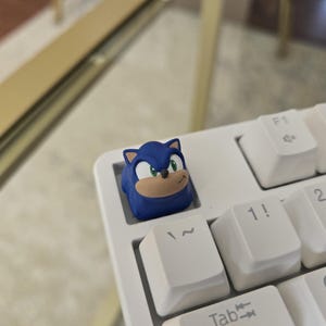 Könnte beinhalten: Ein blauer Sonic the Hedgehog-Tastenkappe ist auf einer weißen mechanischen Tastatur installiert. Die Tastenkappe zeigt das Gesicht der Figur mit grünen Augen und einer hellbraunen Schnauze. Die Tastatur hat weiße Tastenkappen mit schwarzer Beschriftung.