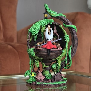 以下が含まれることがあります： ビデオゲーム「Hollow Knight」のキャラクターの精巧なフィギュア。フィギュアは、小さなランタンと赤いマントを着たキャラクターを備えた、木と緑のつるで覆われた構造の中に配置されています。ベースには、追加の小さなフィギュアと葉が含まれています。