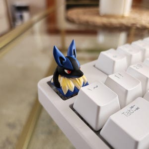 May include: A custom keycap depicting Lucario, in blue, black, and yellow, fitted on a white mechanical keyboard. The keycap is a detailed, artistic representation of the Pokémon character. The keyboard keys are white.