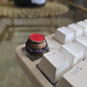 Puede incluir: Una tecla personalizada con un diseño de maceta en miniatura. La tecla tiene un acabado marrón y dorado con una parte superior roja, y se coloca en un teclado blanco. La maceta está decorada con intrincados patrones y una base en forma de dragón.