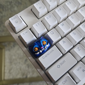 May include: A white mechanical keyboard with a custom blue cat-shaped keycap. The keycap features a detailed design with orange eyes and a menacing grin. The keyboard's keys are arranged in a standard layout.