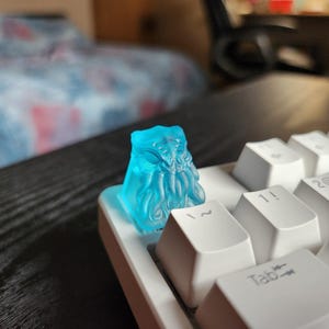 Könnte beinhalten: Ein transluzenter, blau gefärbter Cthulhu-förmiger Keycap, der auf einer weißen mechanischen Tastatur installiert ist. Der Keycap ist mit Tentakeln und Gesichtszügen detailliert. Die Tastatur befindet sich auf einer schwarzen Oberfläche.