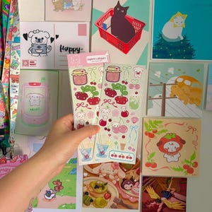 May include: Two vertical sticker sheets featuring a variety of illustrations including red cherries, pink jars, small white cartoon characters, ice cream cones, and blue ribbons on a light green background, held together in a clear plastic sleeve.