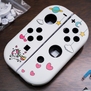 May include: White Nintendo Switch Joy-Con controllers with a unicorn, hearts, clouds, and stars design. The controllers are separated and show the buttons and the back of the controllers.