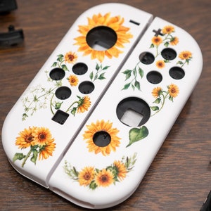 Może przedstawiać: Białe kontrolery Joy-Con do Nintendo Switch z wzorem słonecznika i kwiatów. Kontrolery leżą na drewnianej powierzchni.