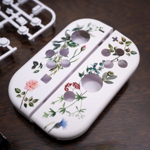 Könnte beinhalten: Weiße Nintendo Switch Konsolenhaut mit einem floralen Design mit Rosen, Blättern und anderen botanischen Elementen.