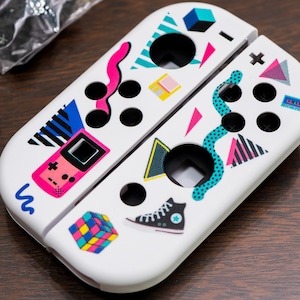 Puede incluir: Mandos Joy-Con blancos para Nintendo Switch con un diseño retro de los años 80 que incluye una Game Boy rosa, un cubo de Rubik, un par de zapatillas Converse y formas geométricas en rosa, azul y verde.