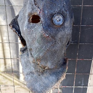 Puede incluir: Una máscara de tela gris oscuro con un solo ojo de botón y un aspecto desgarrado y desgastado. La máscara está hecha de un material grueso y tejido y tiene un agujero para el ojo del usuario.