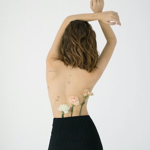 A woman with short brown hair is wearing black trousers and has several small tattoos on her back, including the word "music".  She is holding flowers behind her back.