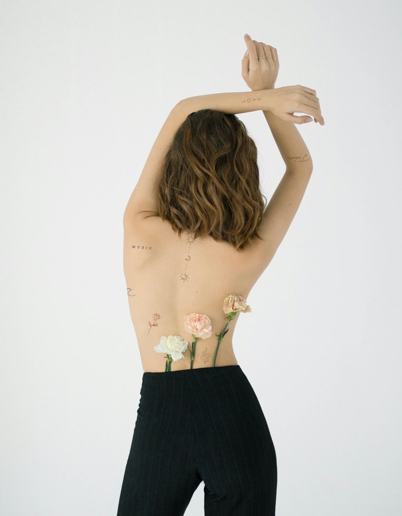 A woman with short brown hair is wearing black trousers and has several small tattoos on her back, including the word "music".  She is holding flowers behind her back.