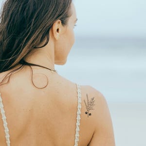 A woman with long brown hair wearing a white crocheted top with a delicate floral paw print tattoo on her shoulder.