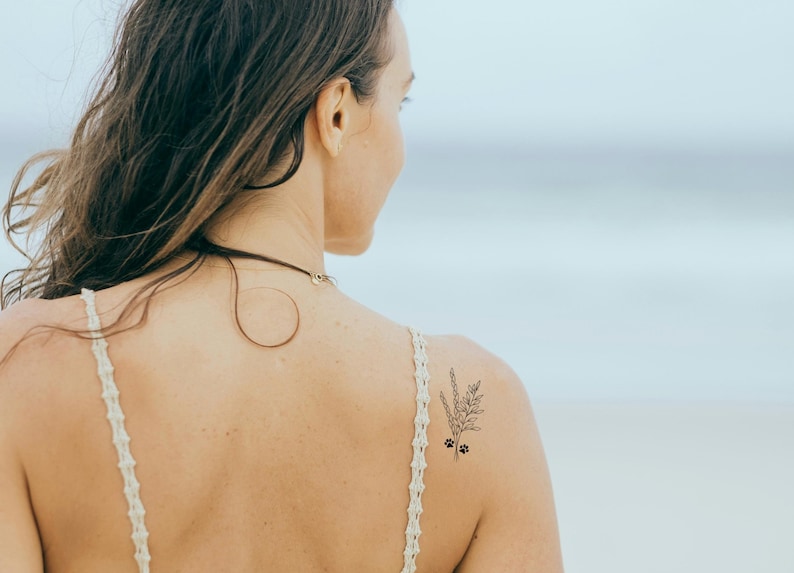 A woman with long brown hair wearing a white crocheted top with a delicate floral paw print tattoo on her shoulder.