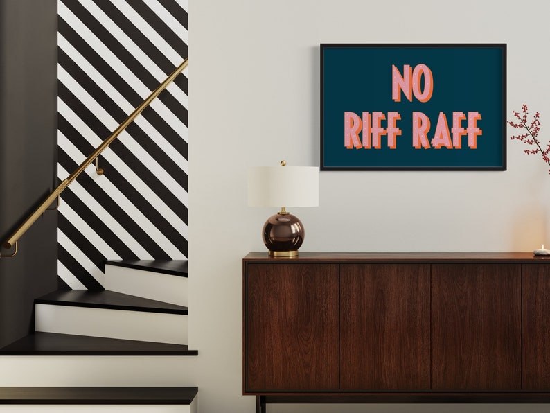 以下が含まれることがあります： 濃い青色の背景に「NO RIFF RAFF」というピンクの文字が書かれた額装されたプリント。プリントは、白い壁に、濃い茶色の木製のキャビネットの上に白いランプが置かれた状態で掛けられています。