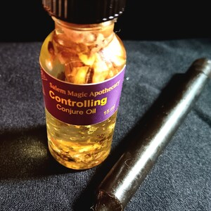 Pode incluir: Uma pequena garrafa de vidro com tampa preta cheia de um l&iacute;quido amarelo e flocos dourados. A garrafa tem um r&oacute;tulo roxo que diz "Salem Magic Apothecary Controlling Conjure Oil 15 ml".