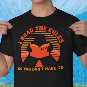 Peut inclure: T-shirt noir avec un graphique de coucher de soleil rétro et le texte "I read the rules so you don't have to". Une silhouette d'une personne lisant un livre avec le mot "rules" sur la couverture est au centre du graphique.