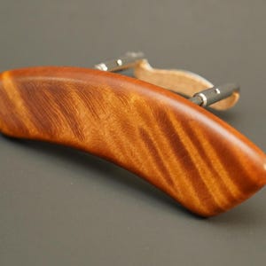 May include: A wooden violin shoulder rest with a natural wood finish and a brown leather strap. The shoulder rest has a curved shape and a smooth surface.