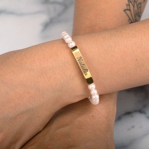 May include: A pearl bracelet with a gold bar engraved with the name "Natalia". The bracelet features small, round, white pearls strung together. The gold bar is rectangular and centered on the bracelet.