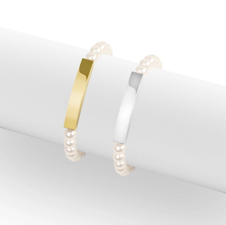 May include: Two bracelets with a pearl bead strand. One bracelet has a gold-colored rectangular bar, and the other has a silver-colored rectangular bar. The bracelets are displayed on a white surface.