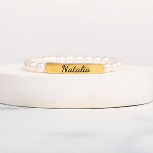 May include: A pearl bracelet with a gold-colored rectangular bar engraved with the name "Natalia" in a script font. The bracelet is displayed on a white, circular pedestal against a white background.