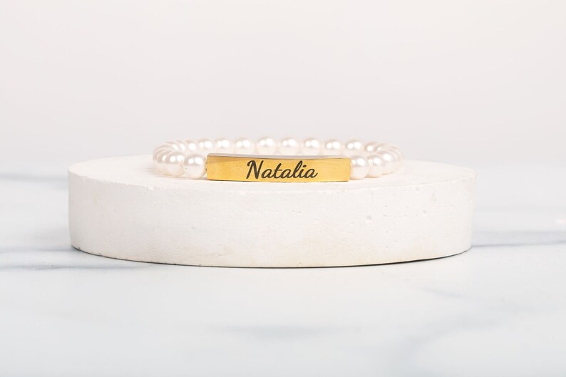 May include: A pearl bracelet with a gold-colored rectangular bar engraved with the name "Natalia" in a script font. The bracelet is displayed on a white, circular pedestal against a white background.