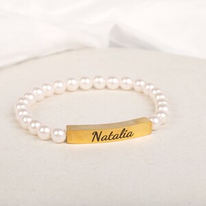May include: A pearl bracelet with a gold-colored bar engraved with the name "Natalia". The bracelet features a string of white, round pearls. The gold bar is curved and centered on the bracelet.