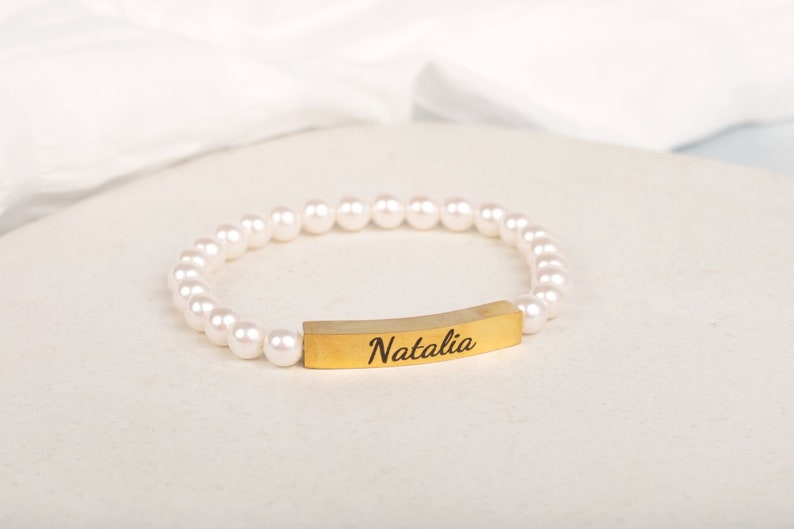May include: A pearl bracelet with a gold-colored bar engraved with the name "Natalia". The bracelet features a string of white, round pearls. The gold bar is curved and centered on the bracelet.
