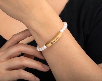 May include: A personalized bracelet featuring a wooden rectangular centerpiece engraved with the name "Natalia." The bracelet is strung with white pearls. The background is a black shirt.