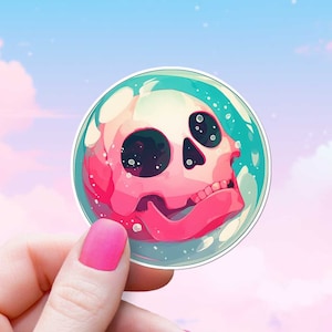 May include: A pink and white skull sticker with a blue and white background. The sticker is in a bubble shape and is being held by a hand.