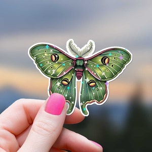 May include: A green and brown luna moth sticker with iridescent glitter accents. The sticker is held in a hand against a blurred background of trees and sky.
