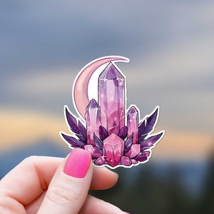May include: A sticker featuring a cluster of pink and purple crystals with a crescent moon behind them. The crystals are surrounded by dark purple leaves. The sticker is held in a hand against a blurred background of a cloudy sky and trees.