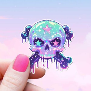 Puede incluir: Una pegatina holográfica de una calavera y huesos cruzados con un diseño de galaxia. La calavera tiene una estrella en la frente y pintura que gotea. La pegatina se sostiene en una mano contra un fondo de cielo rosa y azul pastel.