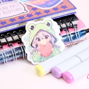 Pode incluir: Um adesivo com uma menina de desenho animado com cabelo rosa usando um moletom verde de sapo e segurando um cora&ccedil;&atilde;o rosa. O adesivo est&aacute; em um caderno com espiral.