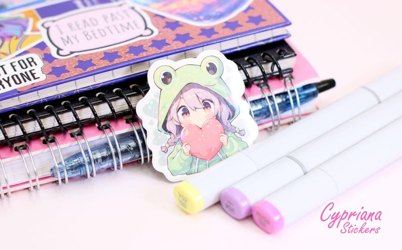 Pode incluir: Um adesivo com uma menina de desenho animado com cabelo rosa usando um moletom verde de sapo e segurando um cora&ccedil;&atilde;o rosa. O adesivo est&aacute; em um caderno com espiral.