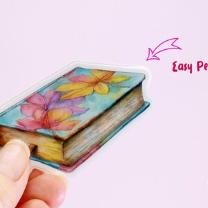 May include: A watercolor sticker of a book with a floral design. The sticker has a clear border and the text "Easy Peel" is visible. The sticker is held by a hand. The sticker is from Cypriana Stickers.