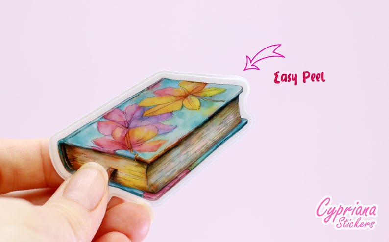 May include: A watercolor sticker of a book with a floral design. The sticker has a clear border and the text "Easy Peel" is visible. The sticker is held by a hand. The sticker is from Cypriana Stickers.