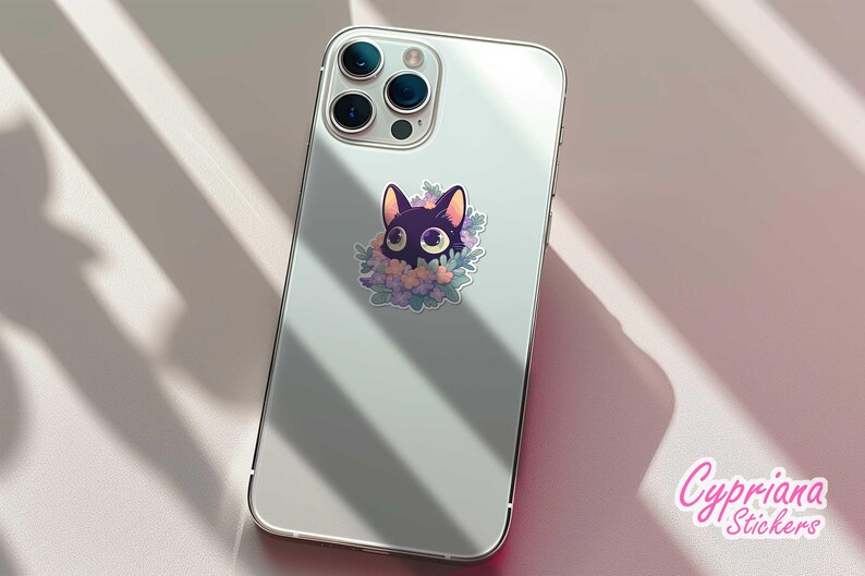 May include: A white iPhone with a black cat sticker on the back. The sticker features a black cat with large eyes and a floral design. The phone is on a white surface.