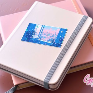 Puede incluir: Una libreta blanca con una pegatina de un horizonte urbano azul y rosa en la portada. La libreta tiene un cierre el&aacute;stico gris. La pegatina tiene una ventana con vista a la ciudad y estrellas.