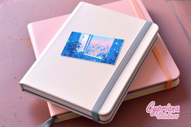 Puede incluir: Una libreta blanca con una pegatina de un horizonte urbano azul y rosa en la portada. La libreta tiene un cierre el&aacute;stico gris. La pegatina tiene una ventana con vista a la ciudad y estrellas.