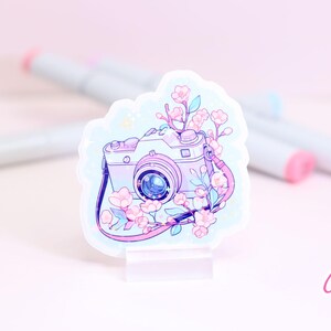 May include: A sticker featuring a pastel-colored camera with a floral design. The camera is light purple with a blue lens and a pink strap. Pink and white flowers and green leaves surround the camera. The sticker is on a clear stand.