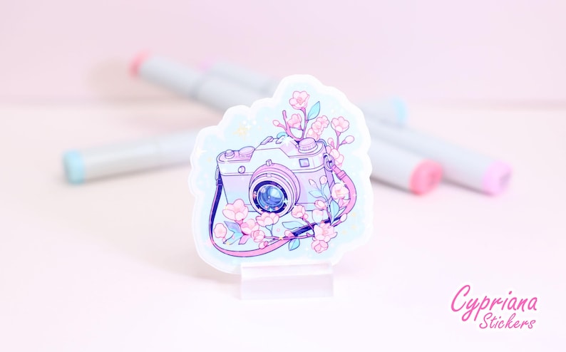 May include: A sticker featuring a pastel-colored camera with a floral design. The camera is light purple with a blue lens and a pink strap. Pink and white flowers and green leaves surround the camera. The sticker is on a clear stand.