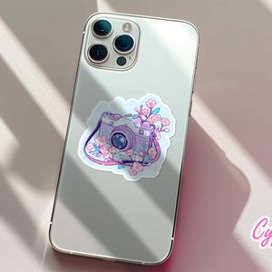 May include: A sticker featuring a vintage camera design with pink and purple accents, adorned with cherry blossoms. The sticker is placed on the back of a silver smartphone. The brand name "Cypriana Stickers" is visible.