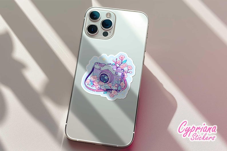 May include: A sticker featuring a vintage camera design with pink and purple accents, adorned with cherry blossoms. The sticker is placed on the back of a silver smartphone. The brand name "Cypriana Stickers" is visible.