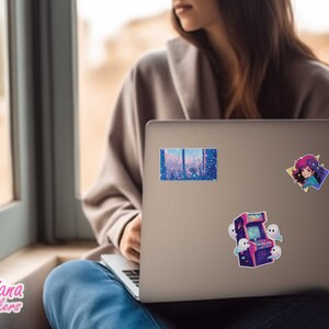 May include: A silver laptop with three stickers on the back. The stickers are of a cityscape, a girl with pink hair, and a retro arcade game with ghosts.
